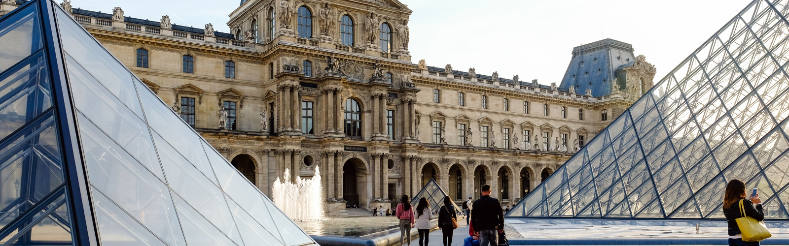 Exterior of the Louvre. The glass pyramid is the focus of the foreground, with the older architecture lining the perimeter. People are walking around the grounds.